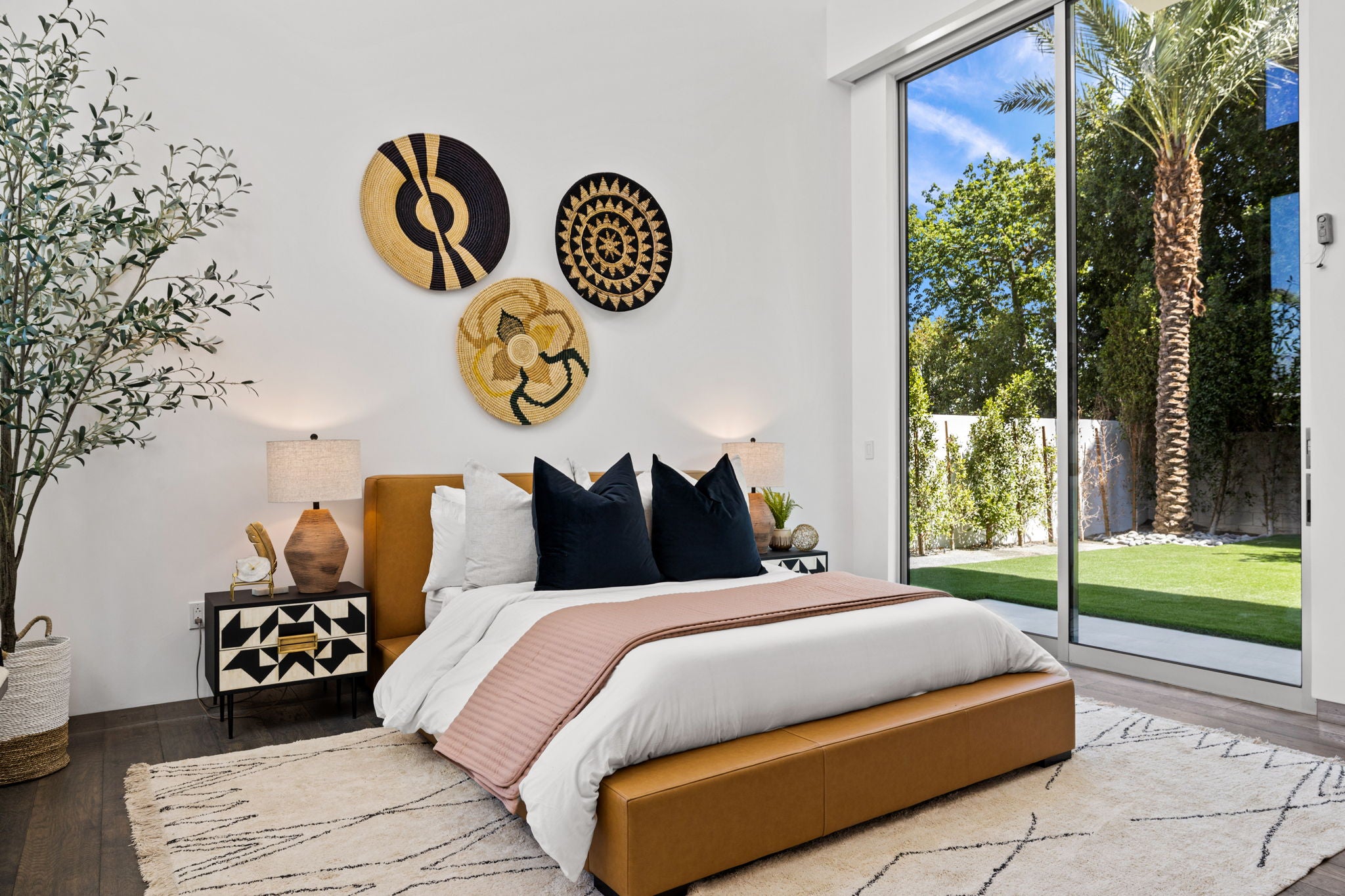 A modern bedroom featuring a low tan leather bed frame with white bedding, a pink throw blanket, and three circular woven wall art pieces. The room has white walls, a patterned rug, and large sliding glass doors looking out to a backyard with a palm tree.