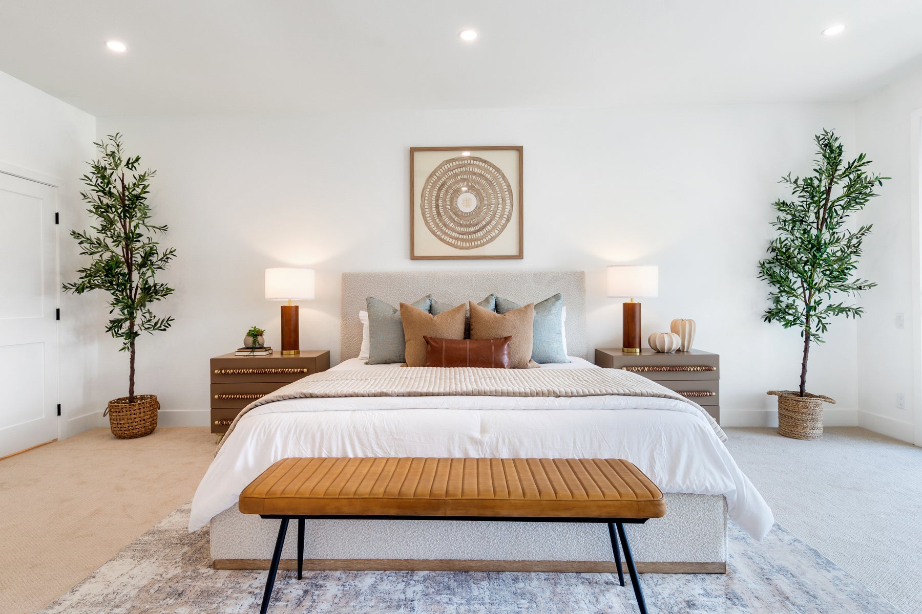 A modern bedroom featuring a bed dressed in white linens with beige and blue-gray pillows, a wooden bench at the foot, matching nightstands and lamps on either side, and two potted trees against a white wall.