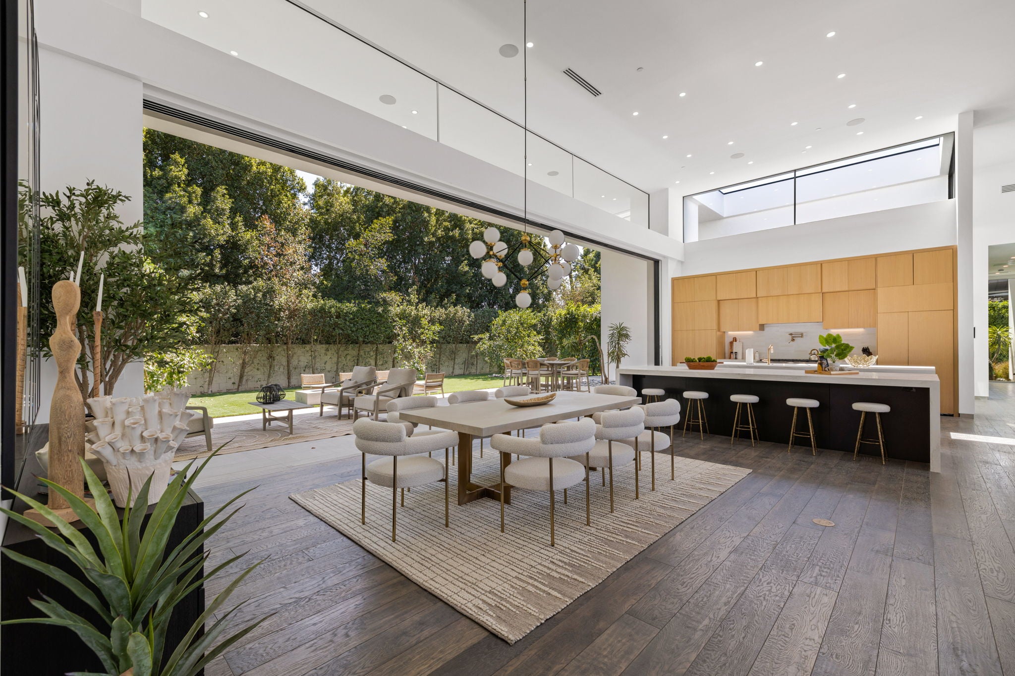 A modern dining room interior with a rectangular light stone dining table, six white upholstered chairs with gold legs, a large woven area rug, dark wood flooring, and an open kitchen with wood cabinetry and a black island. Large glass doors open onto a green outdoor patio and trees.