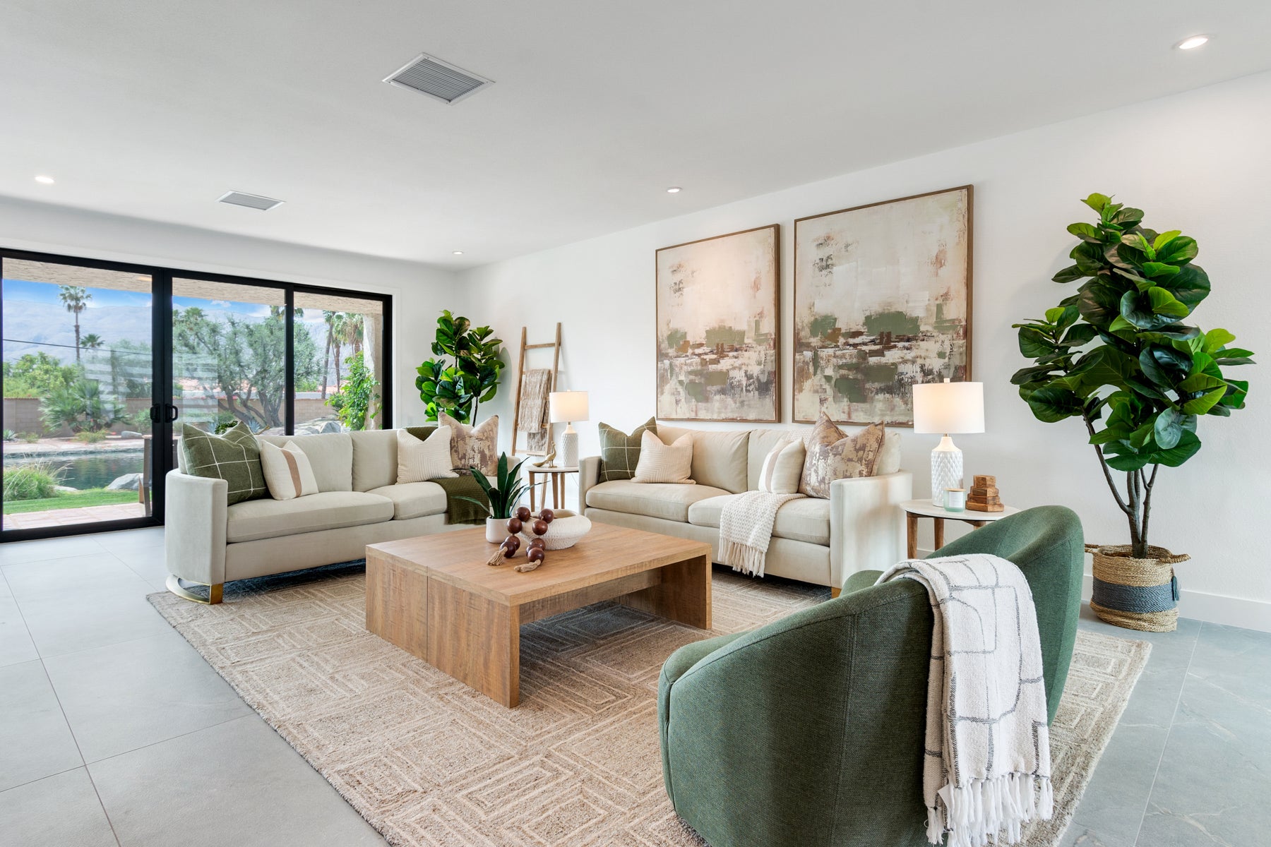 A modern living room featuring a light beige sectional sofa, a green velvet armchair with a white throw blanket, a wooden coffee table, and two large potted fiddle-leaf fig plants. The room has white walls, a light-colored patterned rug, and large windows on the left.