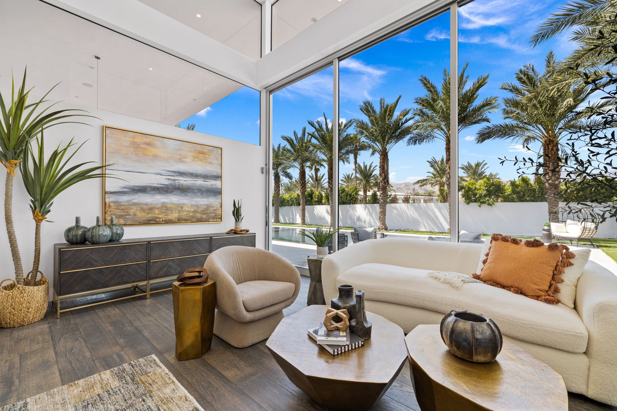 A modern living room interior with a cream curved sofa, matching accent chair, wood coffee tables, a dark gray sideboard, and large floor-to-ceiling windows overlooking palm trees and a blue sky.