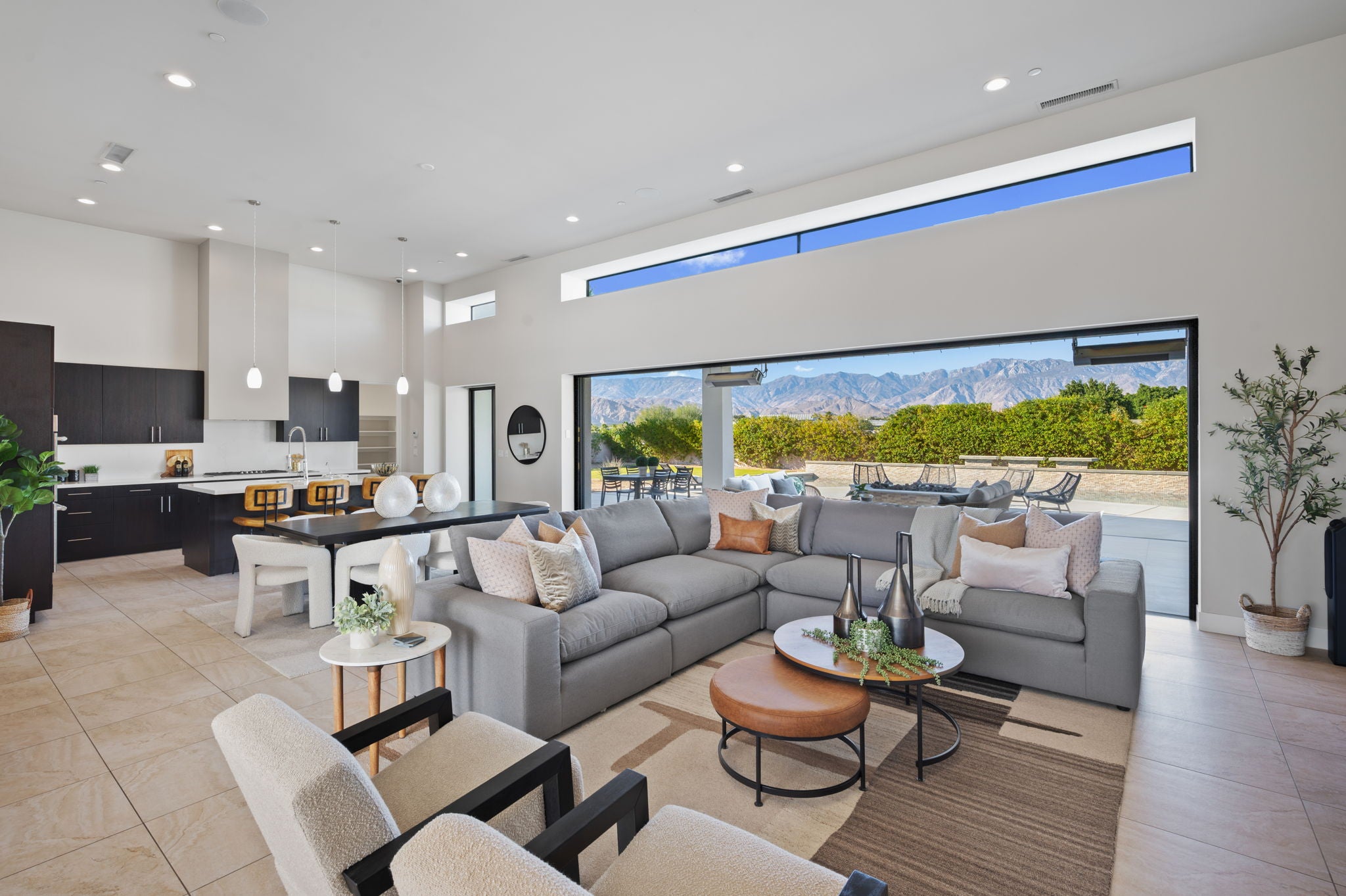 A modern living room featuring a large gray sectional sofa with accent pillows, a round coffee table, and a large floor-to-ceiling window looking out onto a poolside patio and mountains.