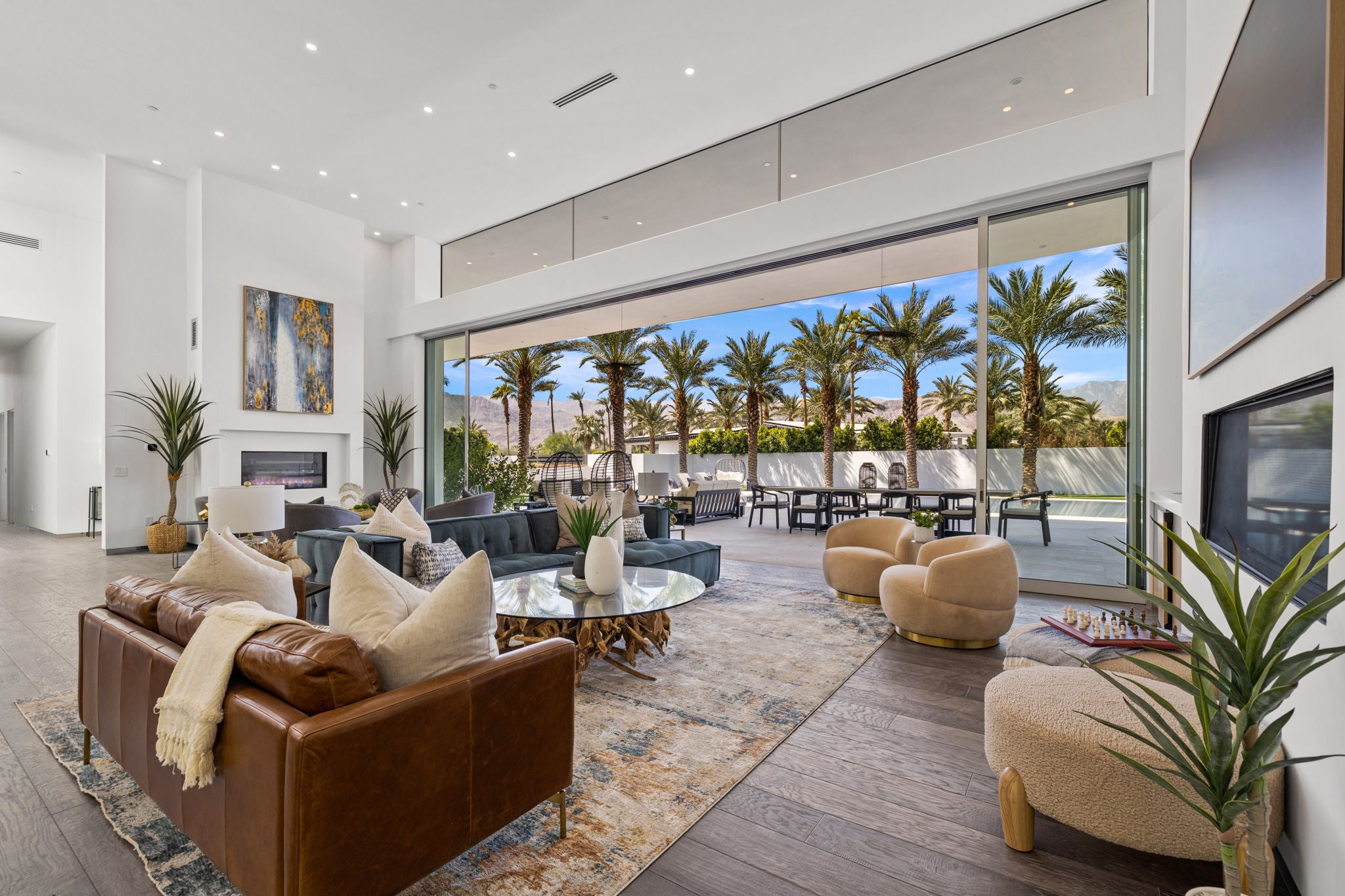 A luxury modern living room with high ceilings, featuring a brown leather sofa, beige armchairs, a glass coffee table, and a large sliding glass door overlooking a poolside patio with palm trees.