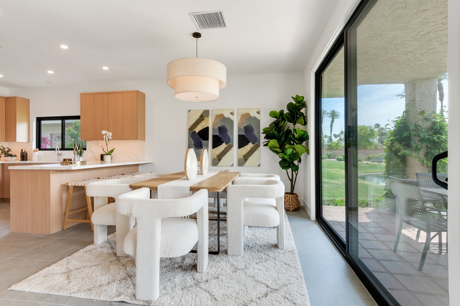 A modern dining room interior with a light wood dining table, white upholstered dining chairs, a white shag rug, and a large potted green plant. The room is connected to a kitchen with light wood cabinets and a white island, with large black-framed sliding glass doors looking out onto a landscaped lawn.