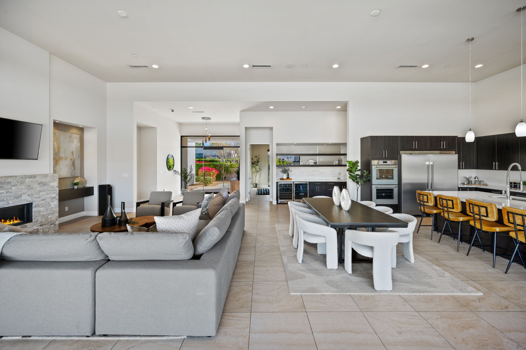 A modern open-plan living room and kitchen featuring a large light gray sectional sofa, a dark wood dining table with white chairs, and a kitchen with dark cabinets and yellow bar stools.