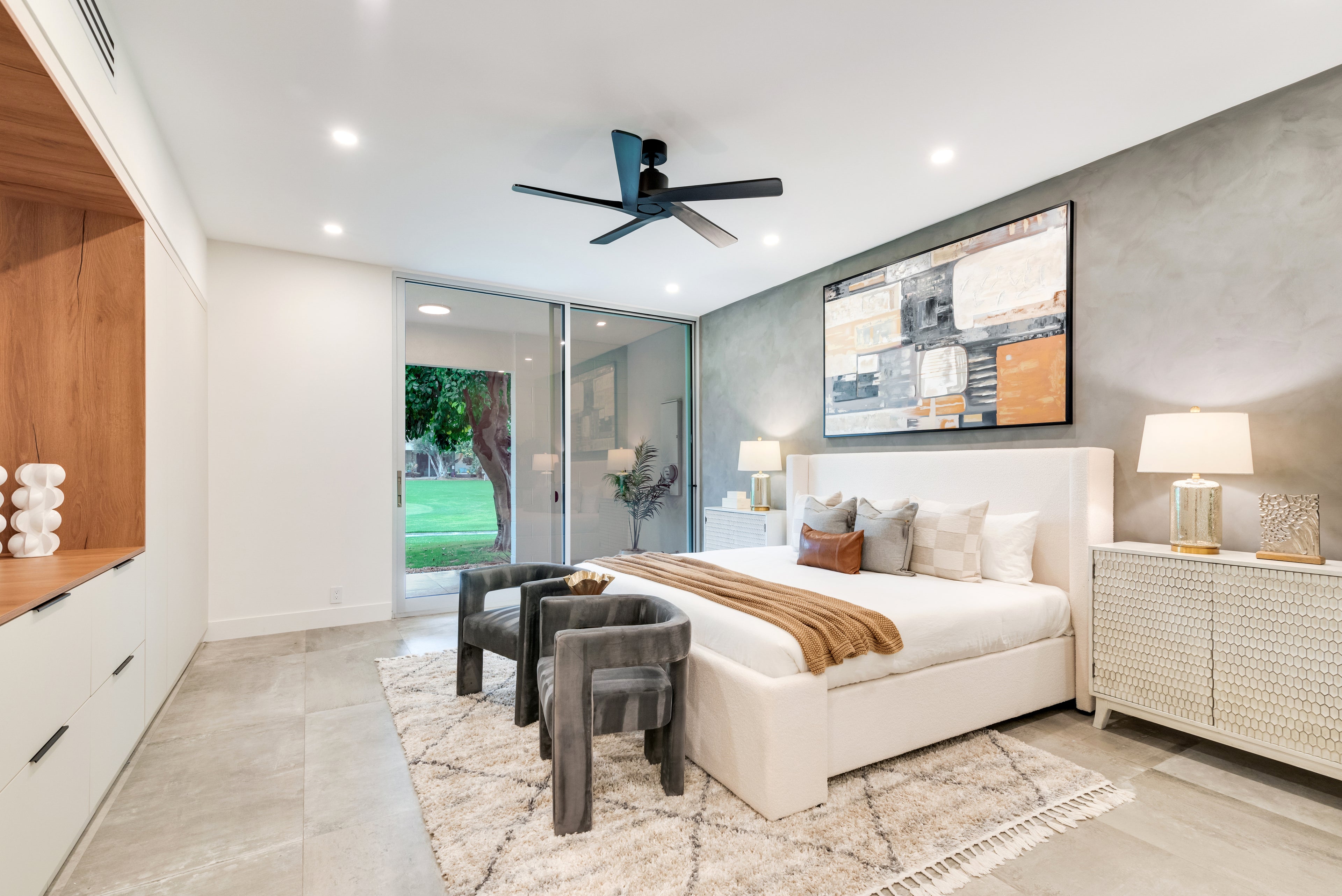 A modern bedroom interior featuring a white platform bed with neutral-toned bedding and a brown throw blanket, set against a textured gray accent wall with a large framed artwork. The room includes a white nightstand with a lamp, a light wood cabinet with open shelving, a patterned rug, two dark gray chairs, and a ceiling fan.