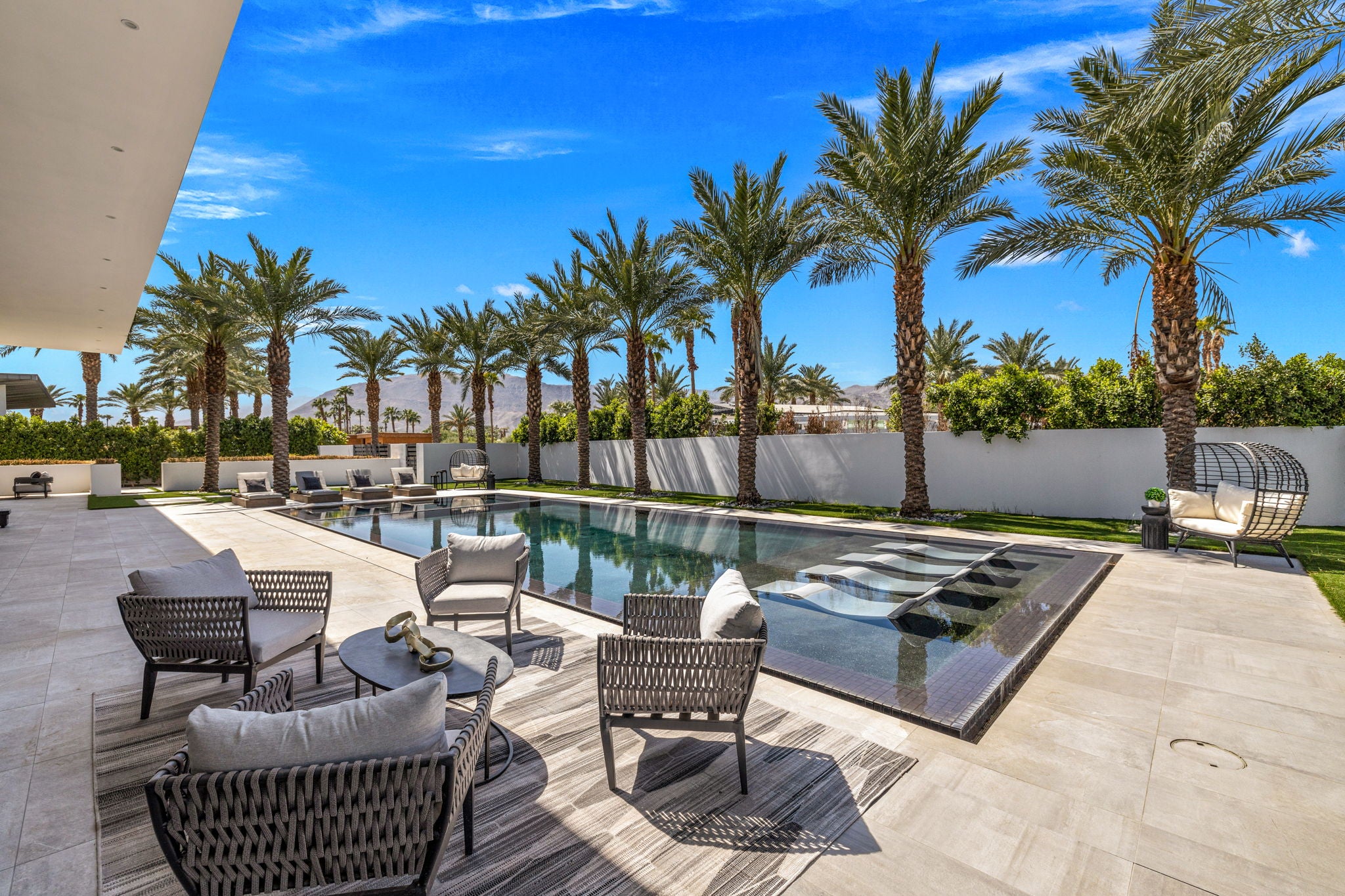 Modern outdoor patio area with a rectangular swimming pool, light stone tile deck, palm trees, and outdoor seating including woven chairs, tables, and a hanging lounge chair under a bright blue sky.