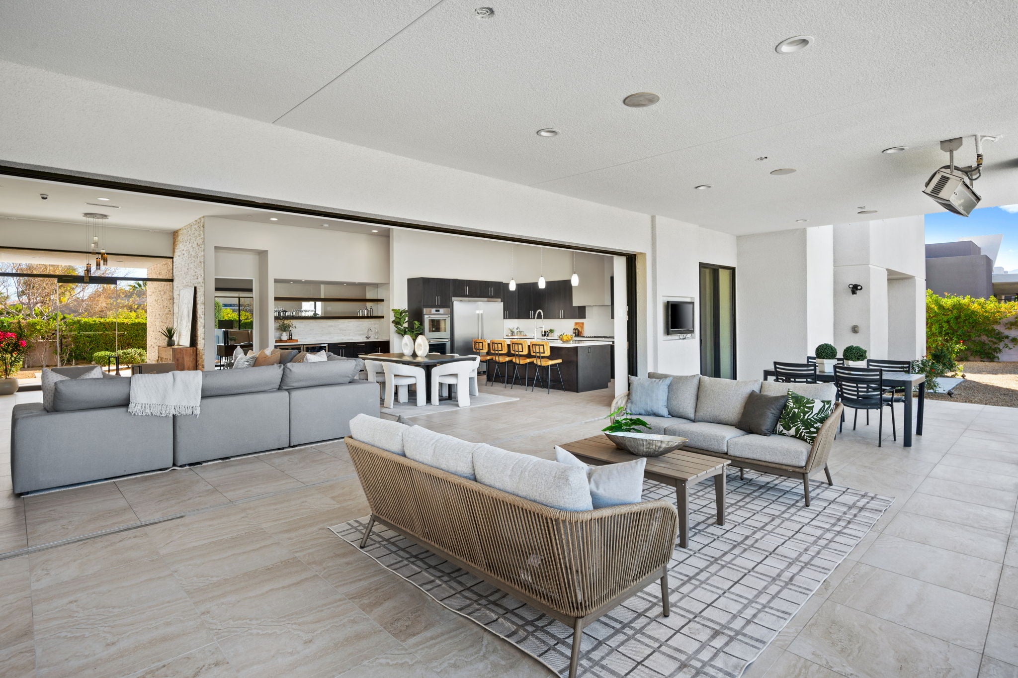 A modern outdoor patio living room featuring a woven wicker sofa with white cushions, a large gray sectional sofa, a patterned tile rug, and an open kitchen in the background.