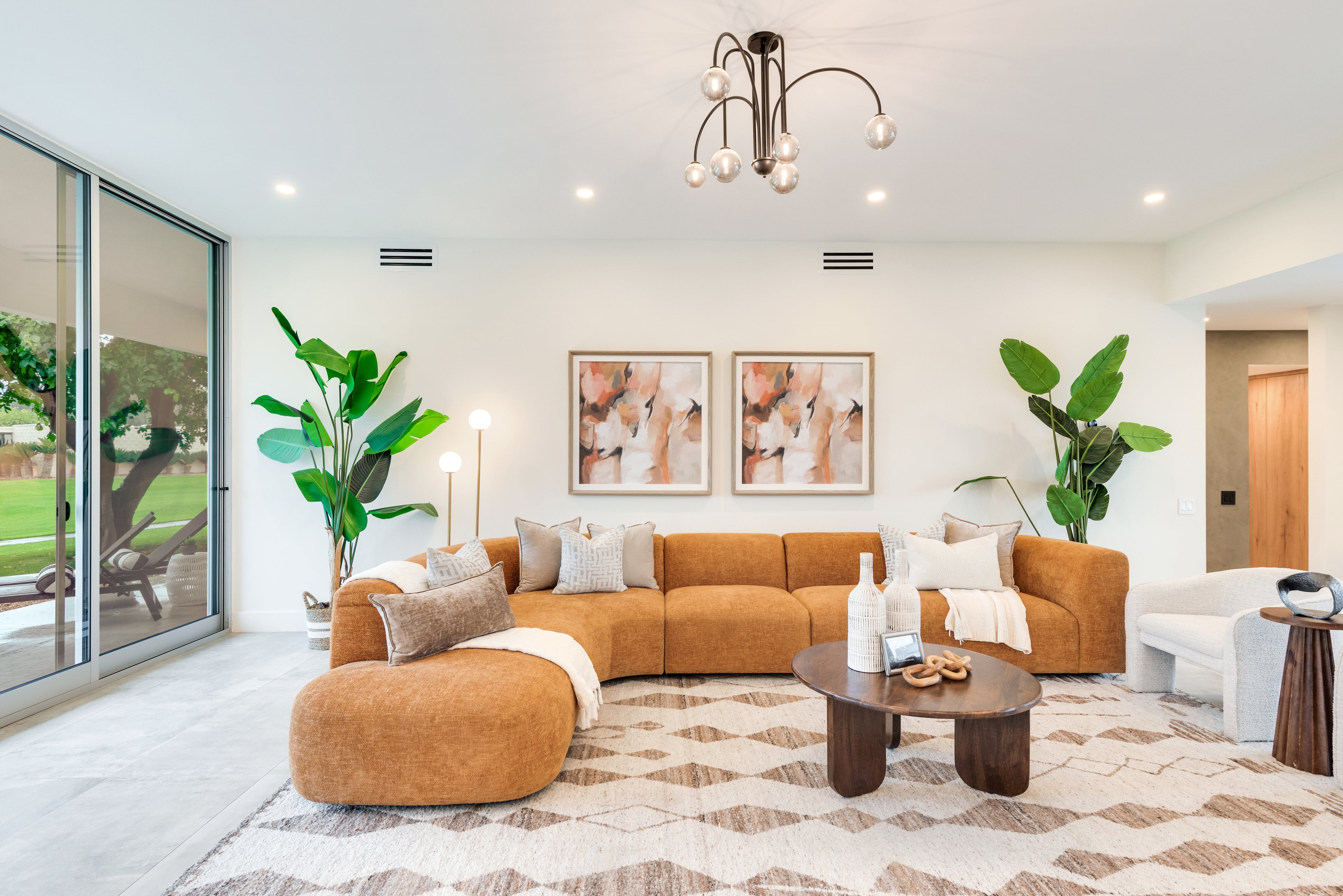 A modern living room featuring a curved rust-orange sectional sofa with matching ottoman, styled with neutral throw pillows, a patterned area rug, a dark wood coffee table, and flanking green plants.