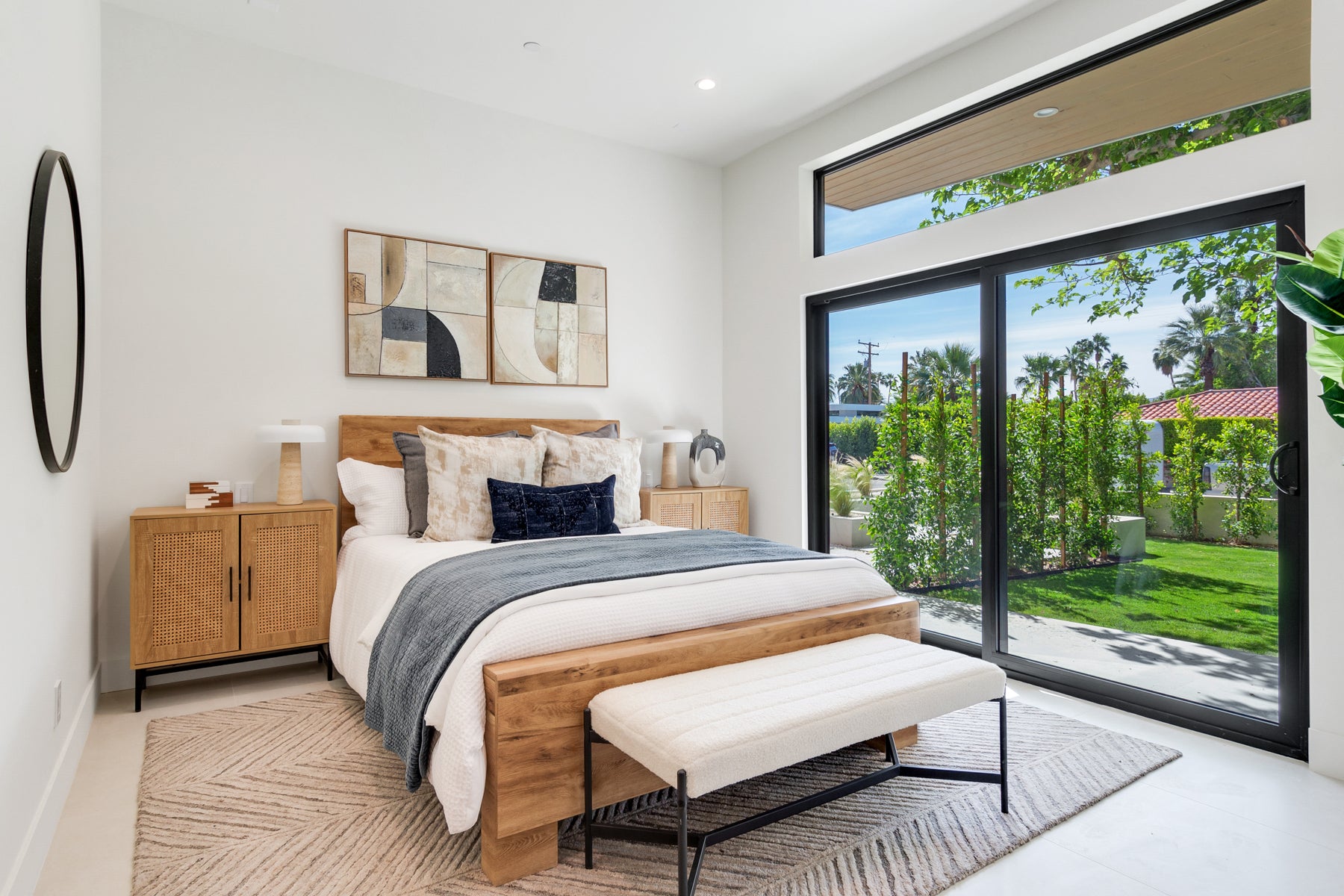 A modern bedroom featuring a wooden bed frame with white bedding, a gray throw blanket, a matching wooden nightstand, and a cream upholstered bench at the foot of the bed. The room has white walls, a patterned beige rug, and large black-framed sliding glass doors looking out onto a green backyard.