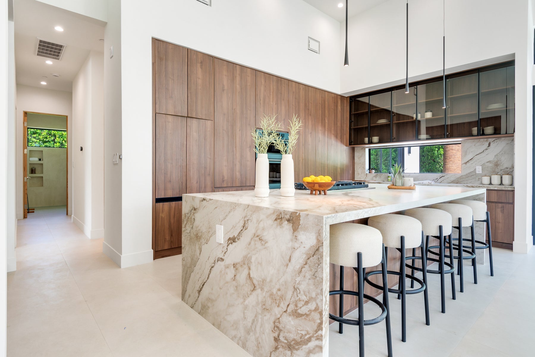 A modern kitchen island with a beige and white marble-patterned countertop and matching marble base, surrounded by white bar stools with black metal frames. The background features dark wood cabinetry, glass-front upper cabinets, and a window looking out onto greenery.