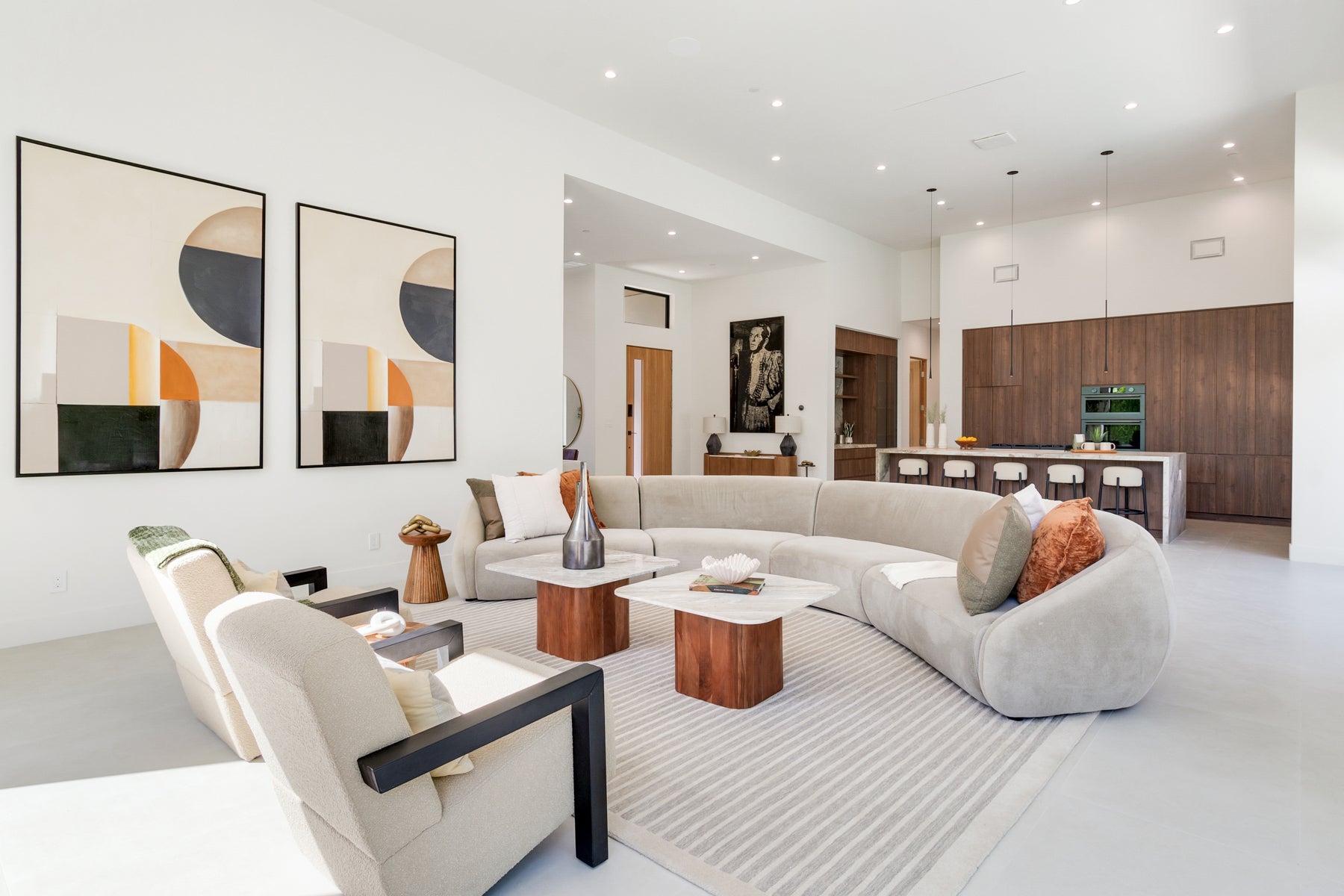 A modern living room featuring a large curved light gray sectional sofa, two matching beige armchairs, low wooden coffee tables, a striped area rug, and two large abstract wall art prints on the white wall. Palm Springs, CA. 