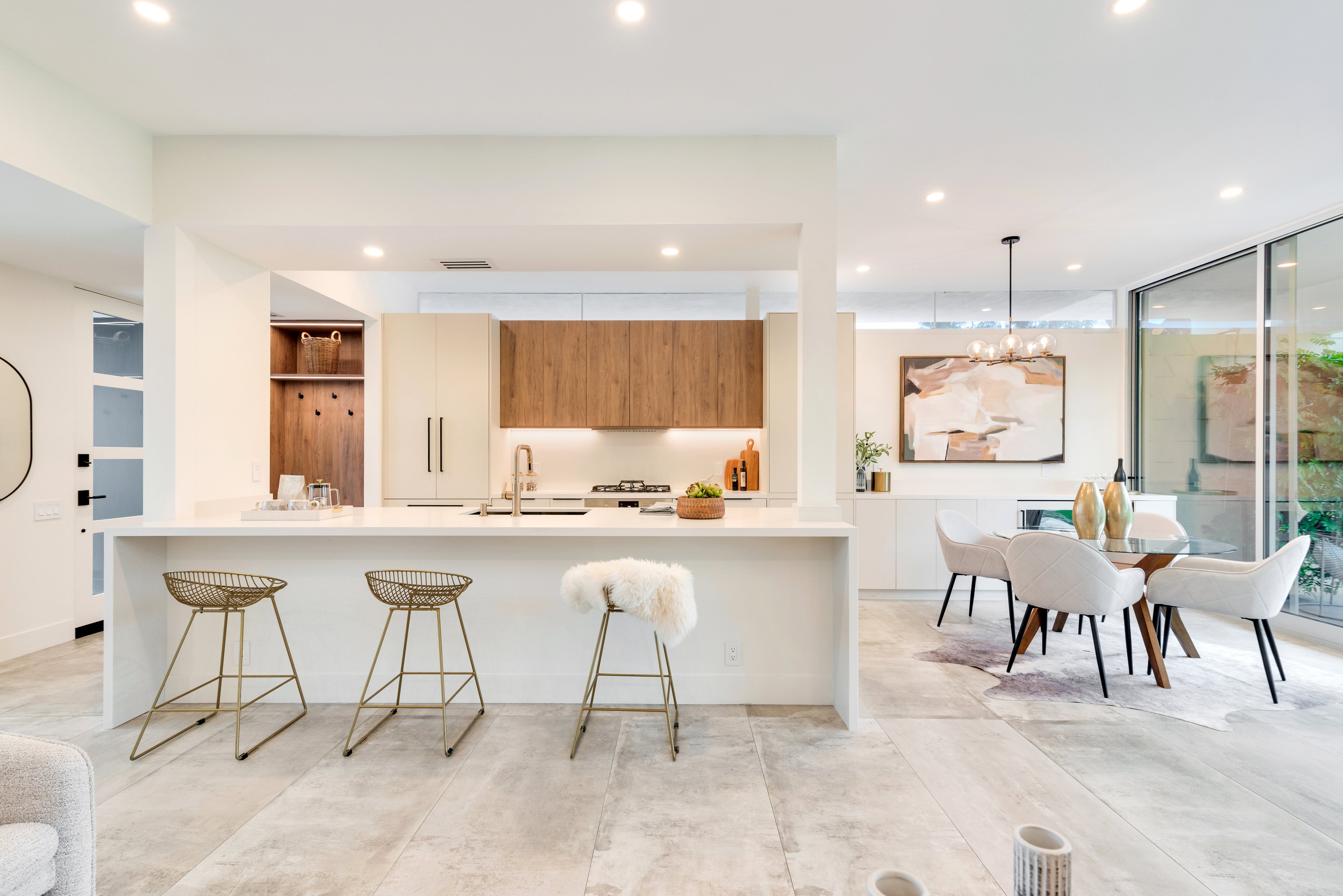 A modern kitchen island with a white countertop and backsplash, three wire bar stools, and a dining area with white chairs and a wooden table in an open-plan interior.